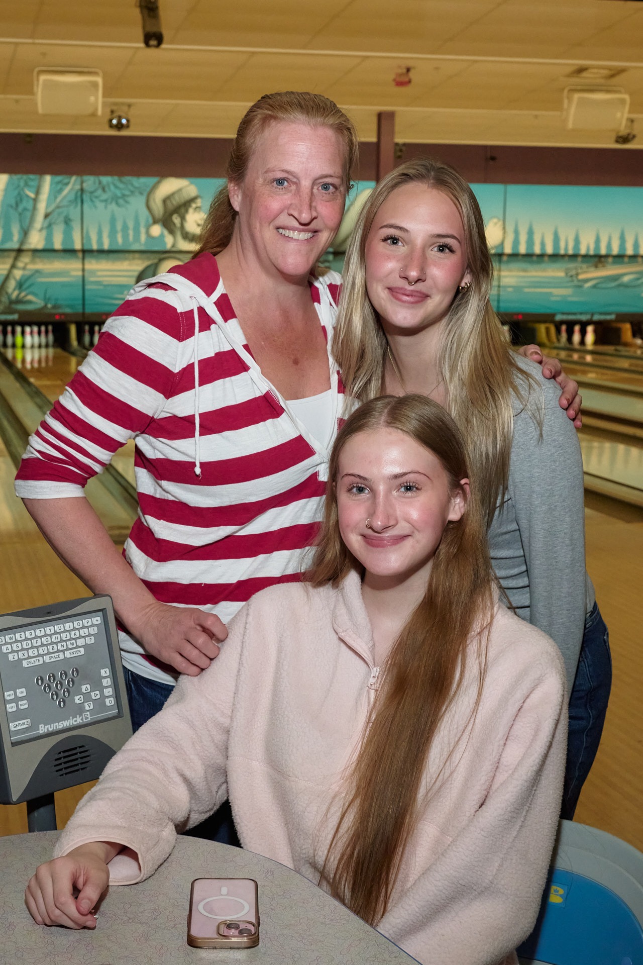 A smiling mother and her two teenage daughters posing together at a bowling alley.