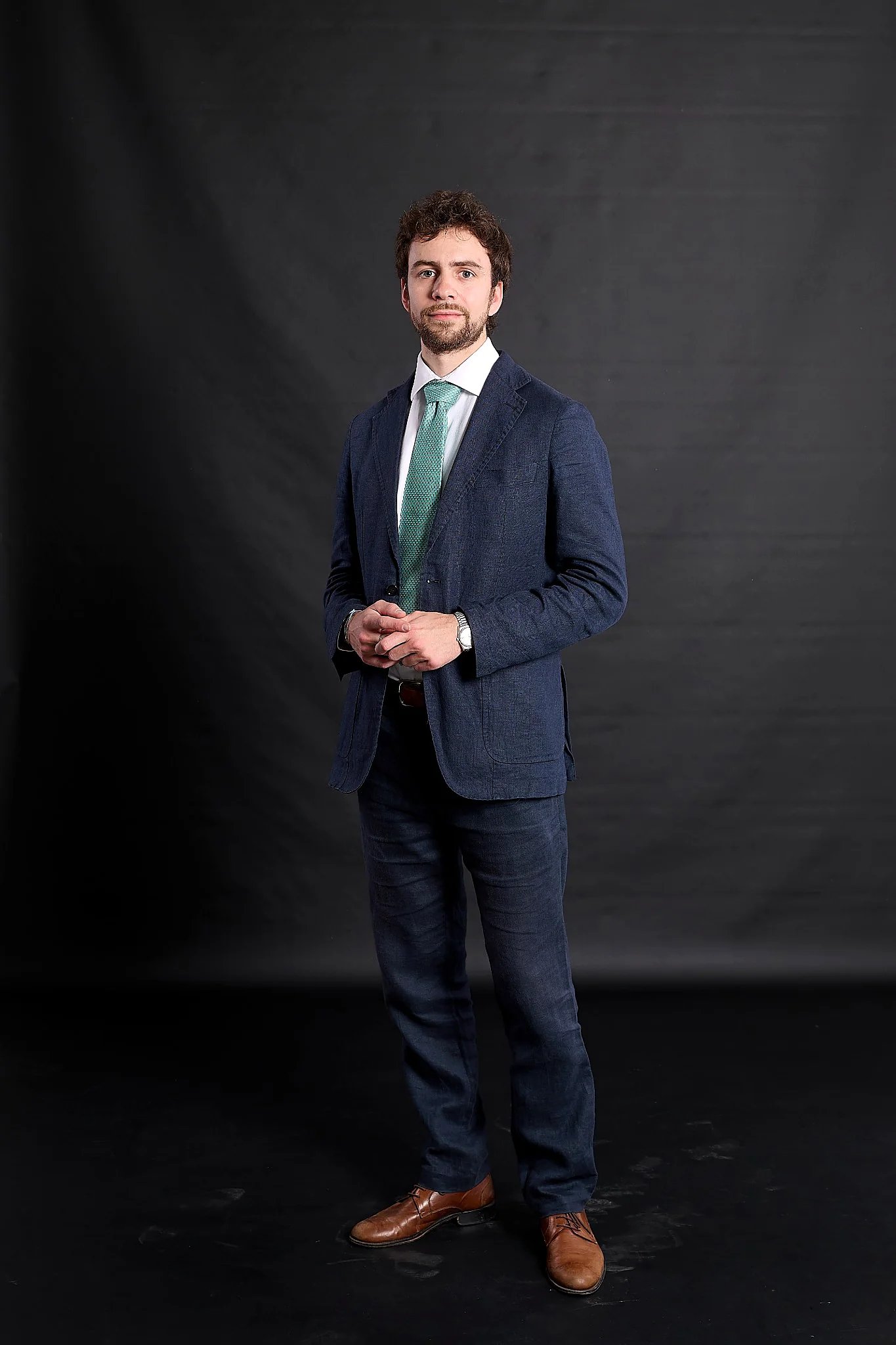 Professional man in a blue textured suit and green tie posing against a black background.
