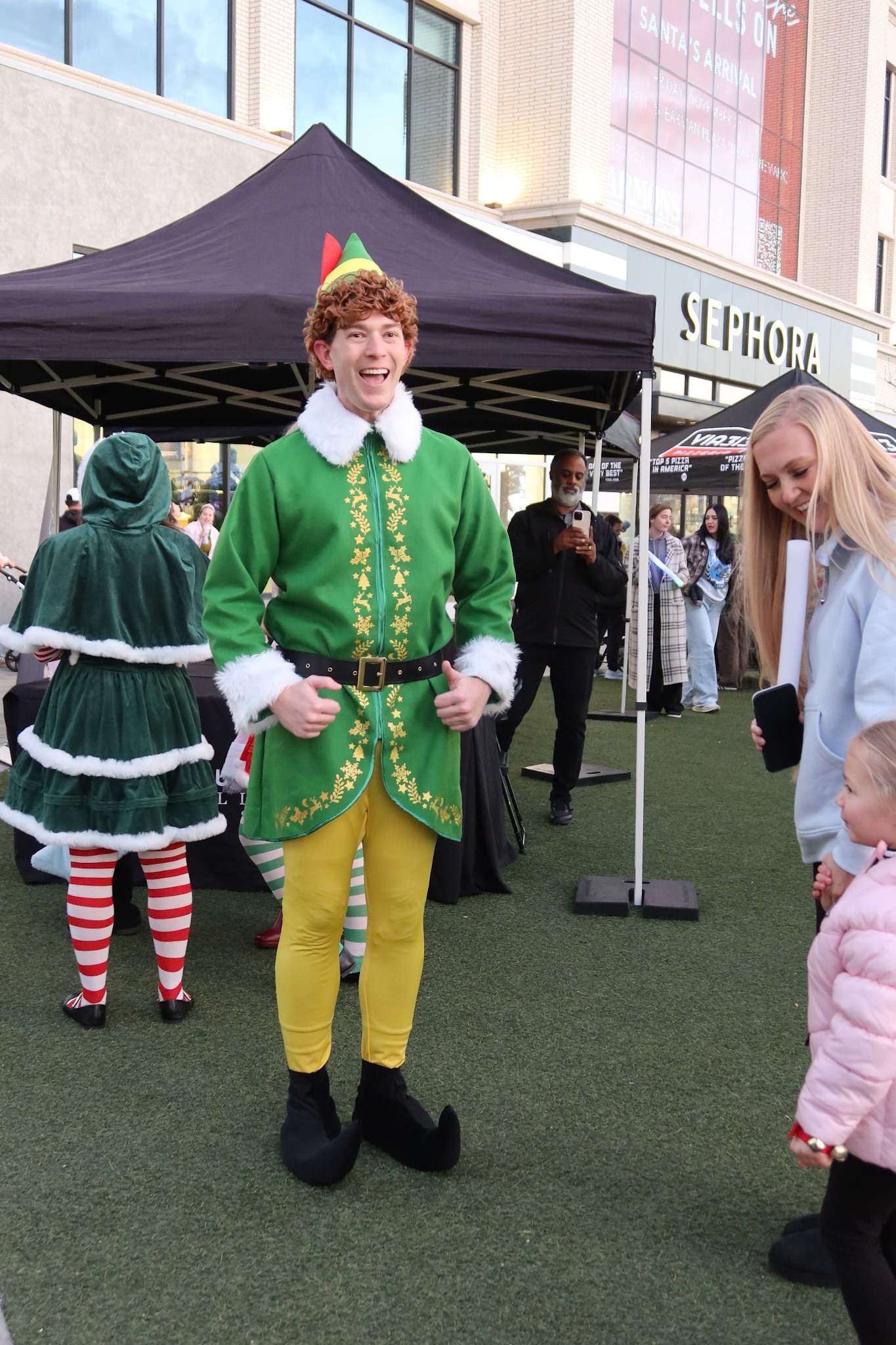 Man dressed in a Buddy the Elf costume gives a thumbs up during a festive holiday event at an outdoor mall.