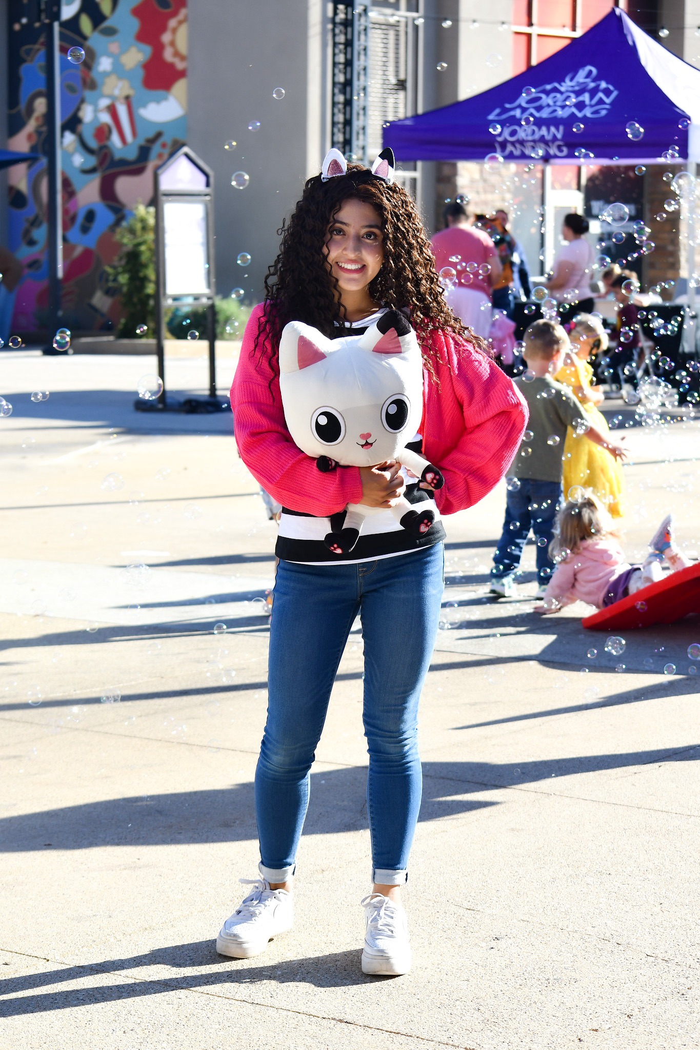 A girl in a pink sweater holds a Gabby's Dollhouse Pandy Paws plush amid floating bubbles at Jordan Landing.