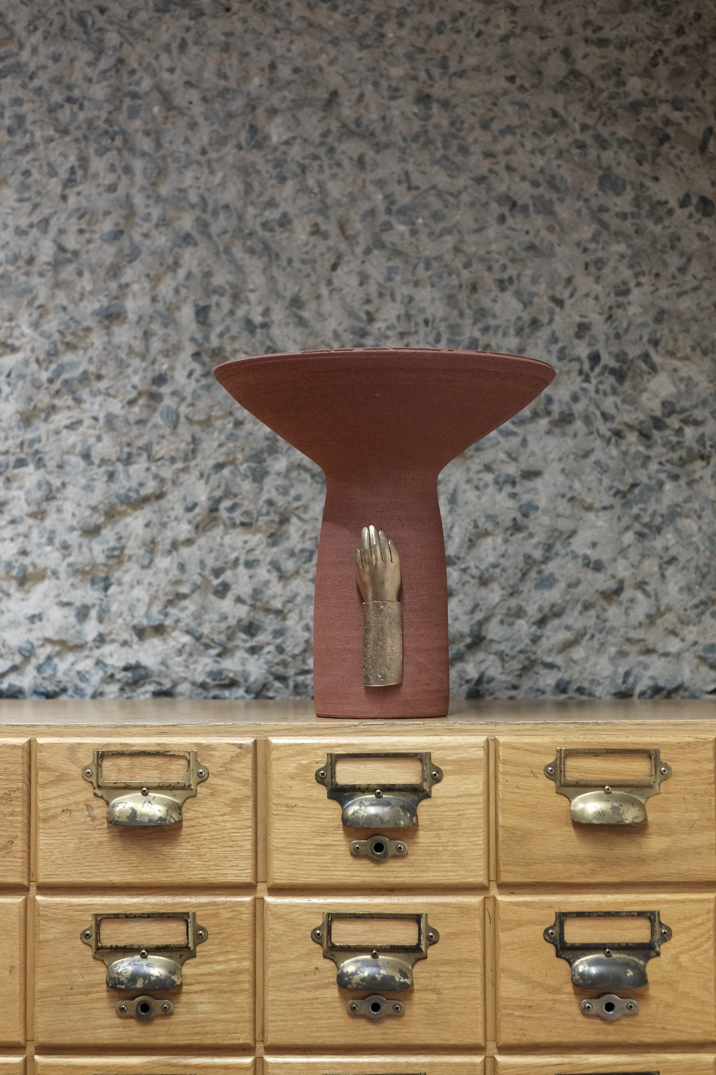 Tall ceramic vessel in deep red stoneware, with a bronze sculpted hand mounted on the base.