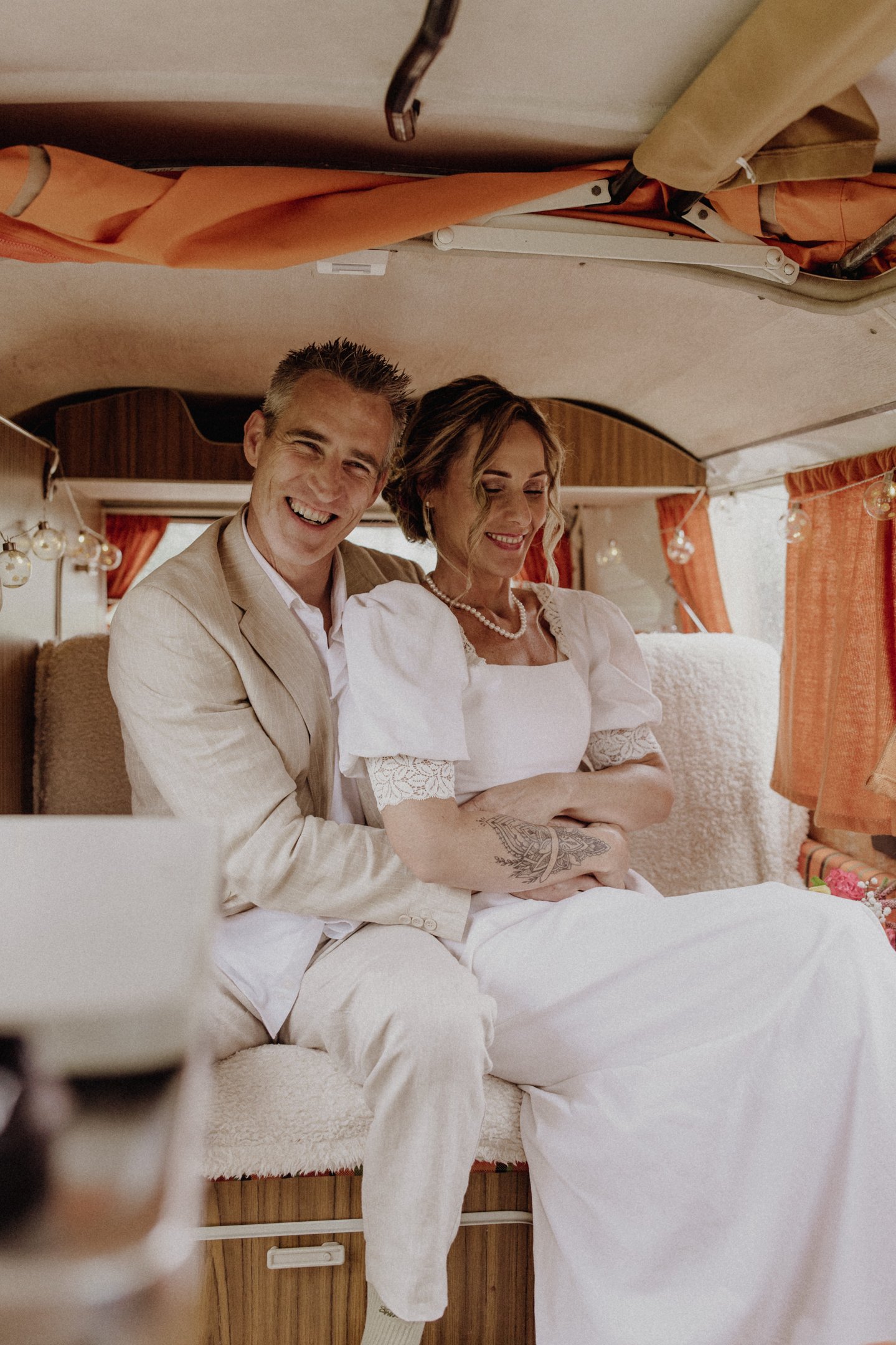 Smiling bride and groom sitting inside a vintage camper van after their wedding ceremony.