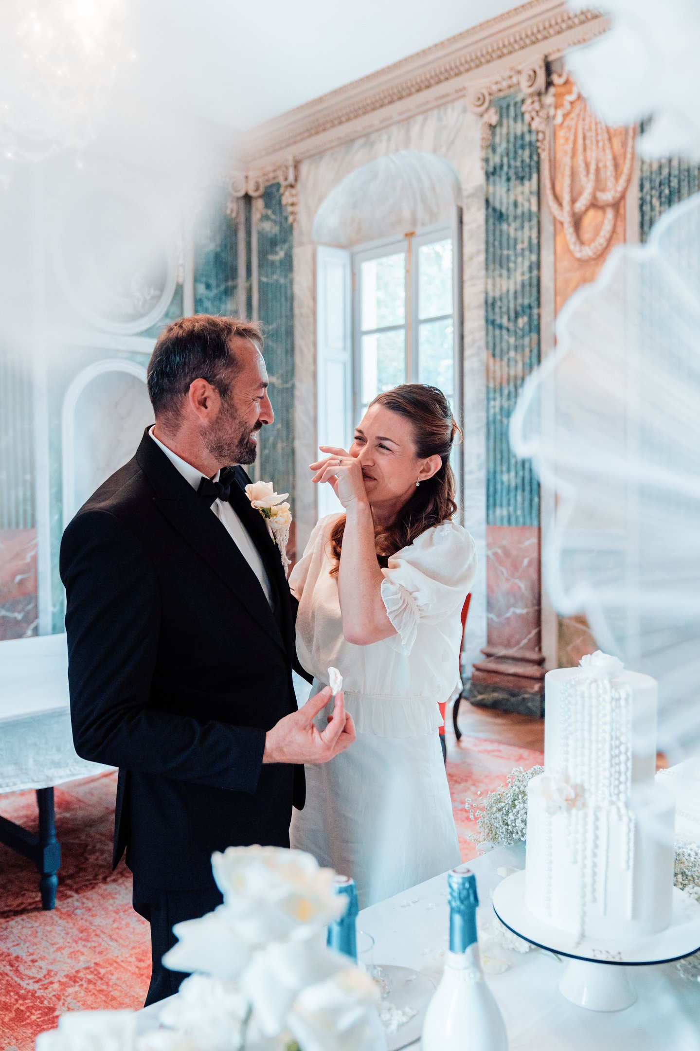 A smiling bride and groom share a joyful moment by their tiered white wedding cake in a luxury venue.