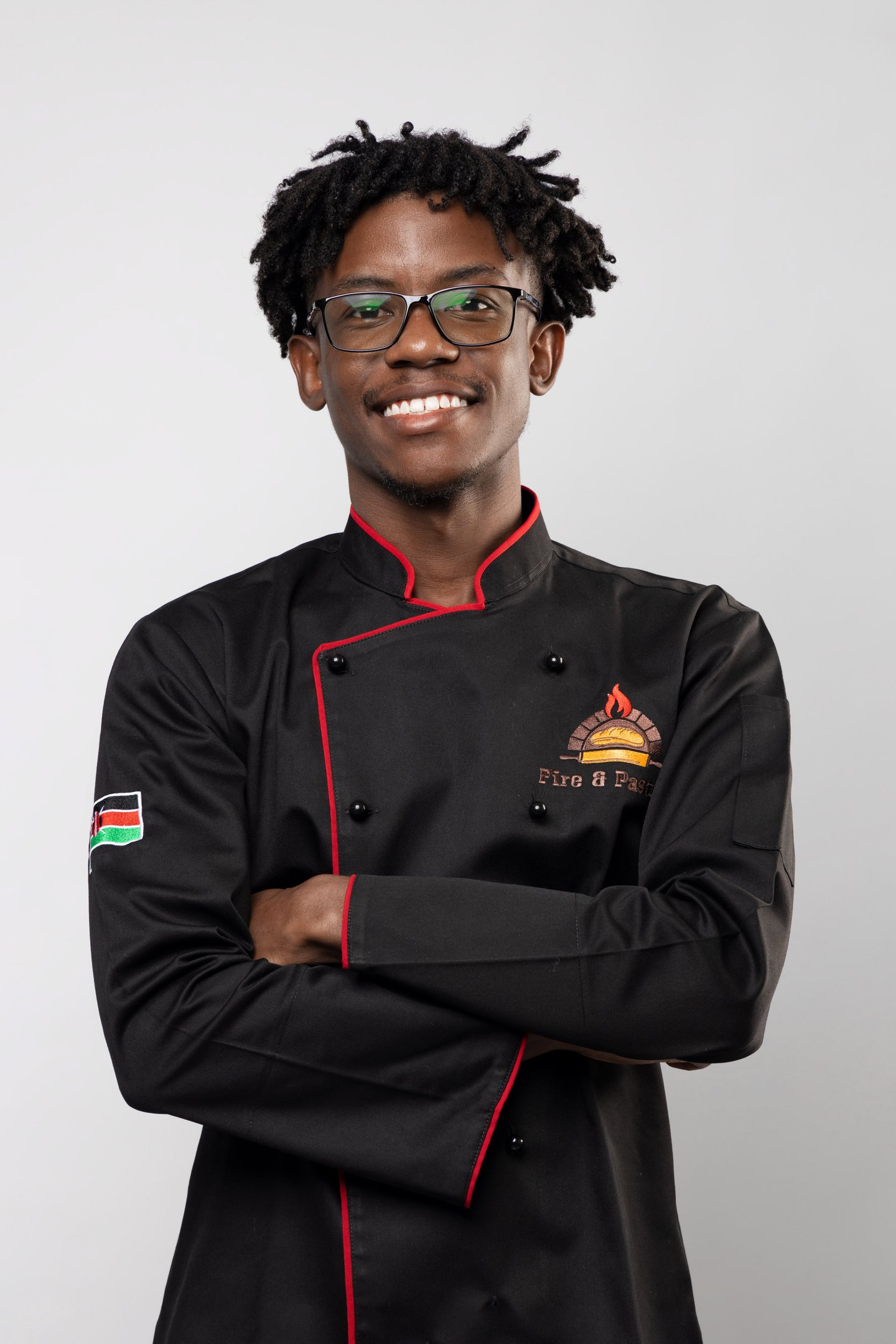 A smiling young chef wearing a professional black double-breasted culinary jacket with red trim.