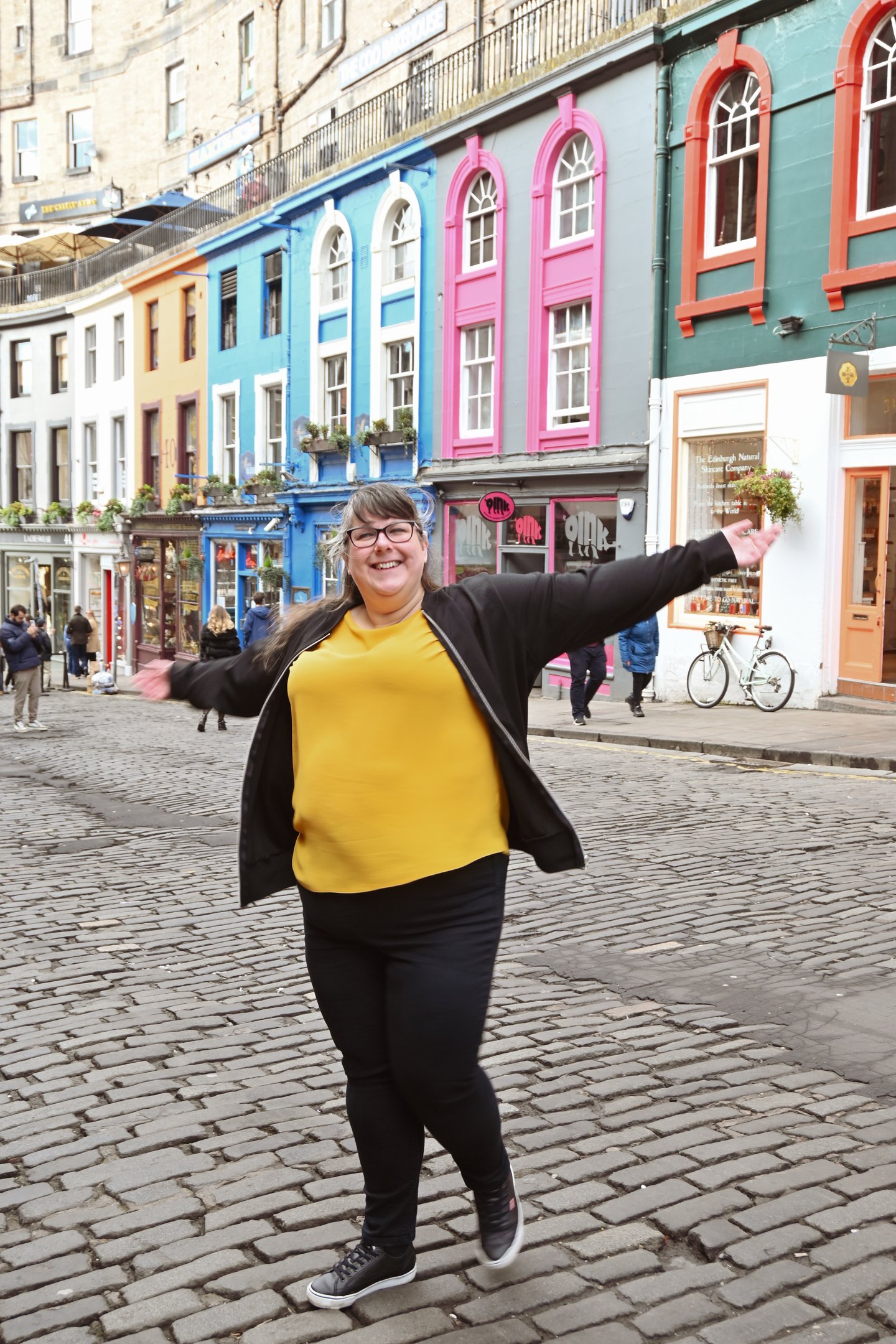 Laura is dancing with her arms out on a cobbled street with brightly coloured buildings behind her