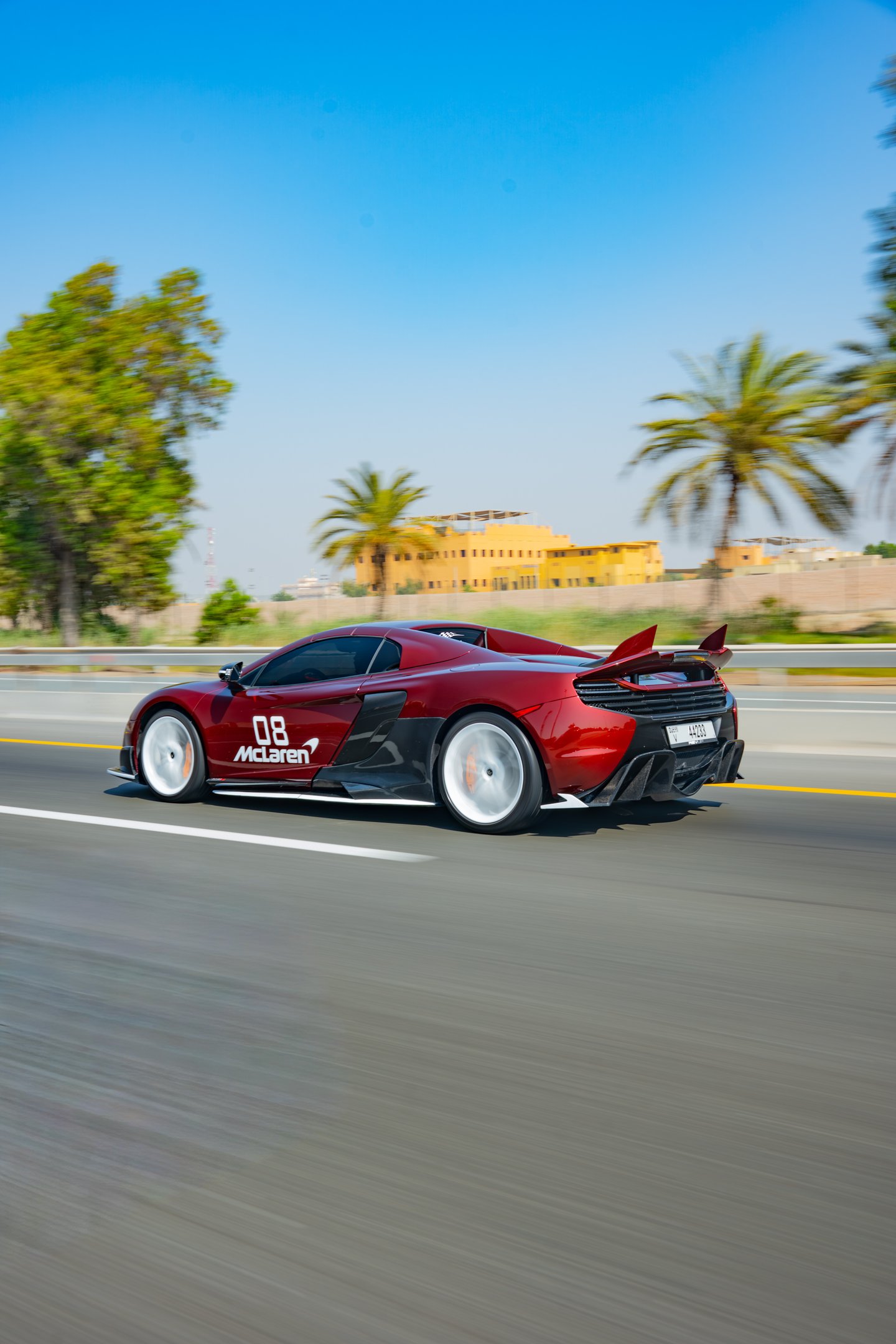 A red McLaren 650S Spider luxury supercar driving fast on a highway with palm trees.