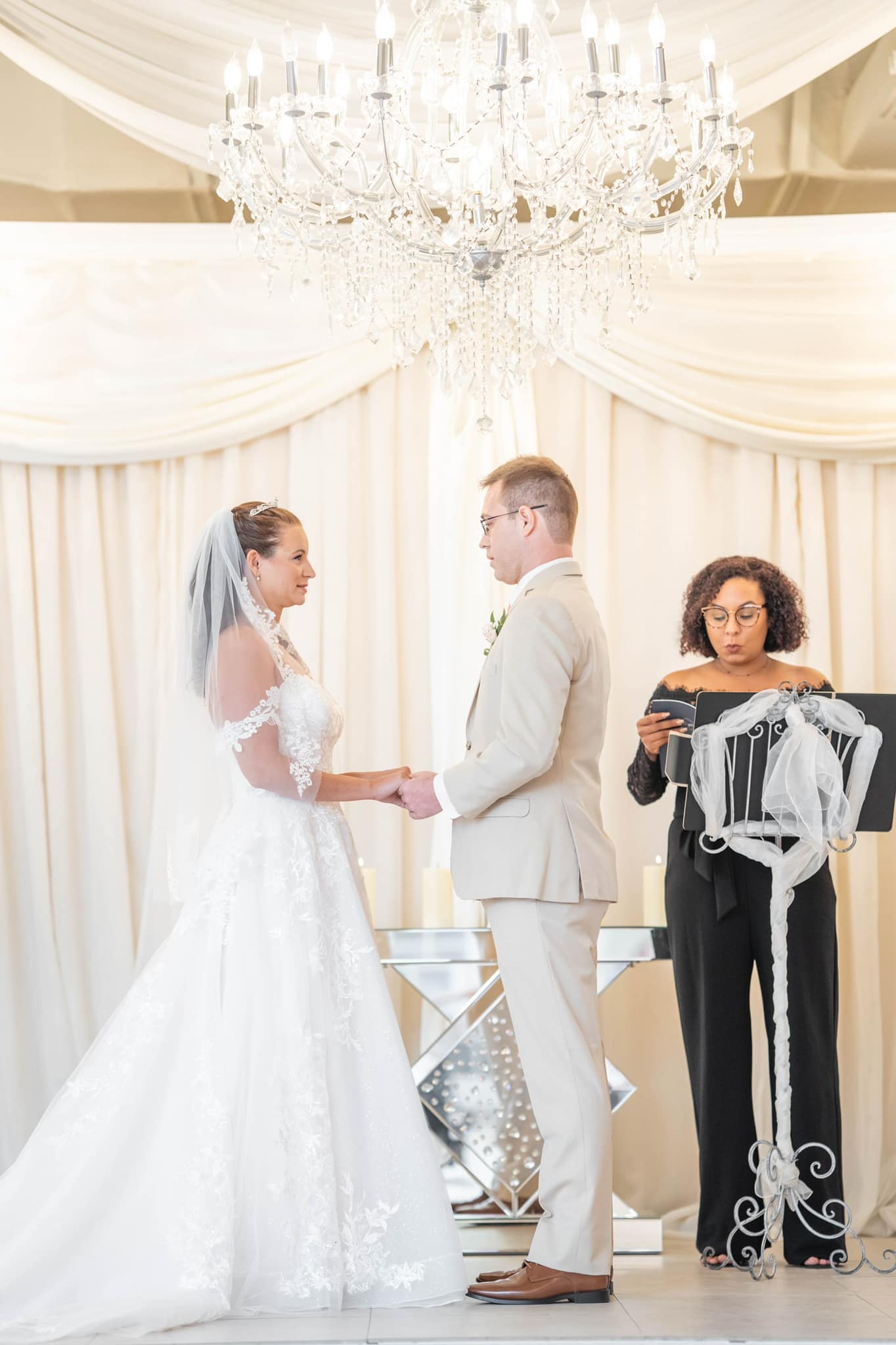 Bride and groom exchanging wedding vows under a crystal chandelier with an officiant.