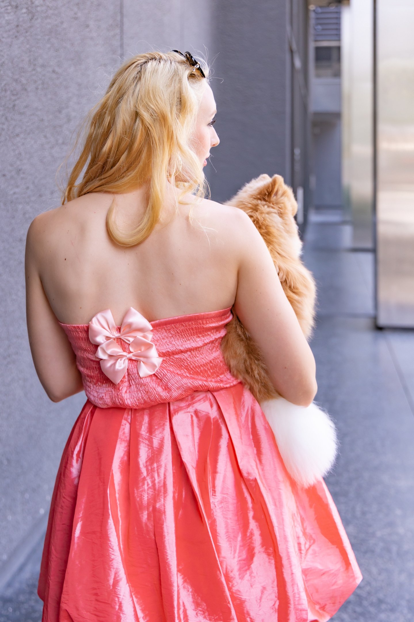 An editorial-style shot from behind of a blonde woman in a coral dress holding a toy Pomeranian