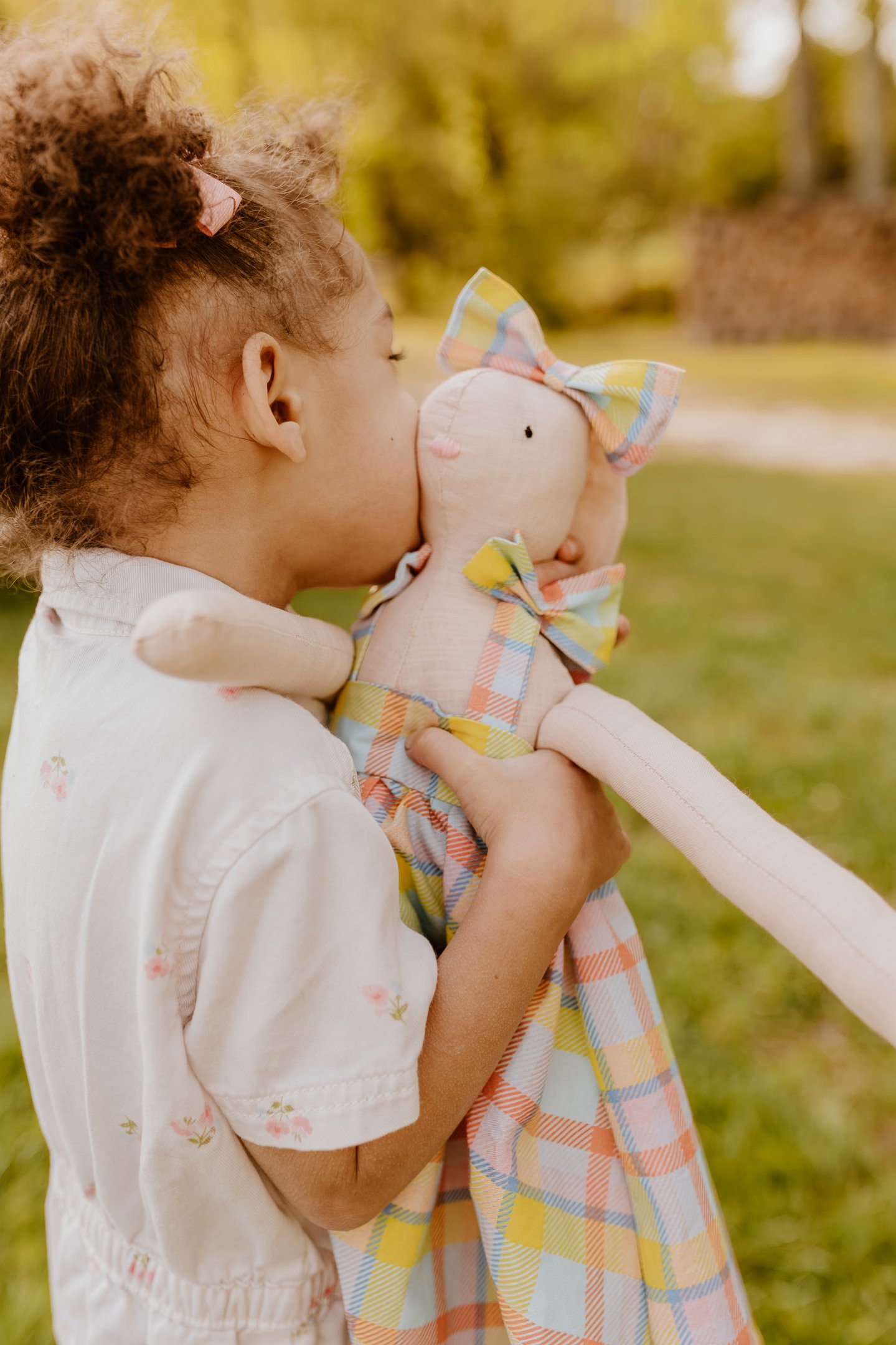 A young girl with curly hair hugs and kisses a handmade linen bunny doll outdoors.