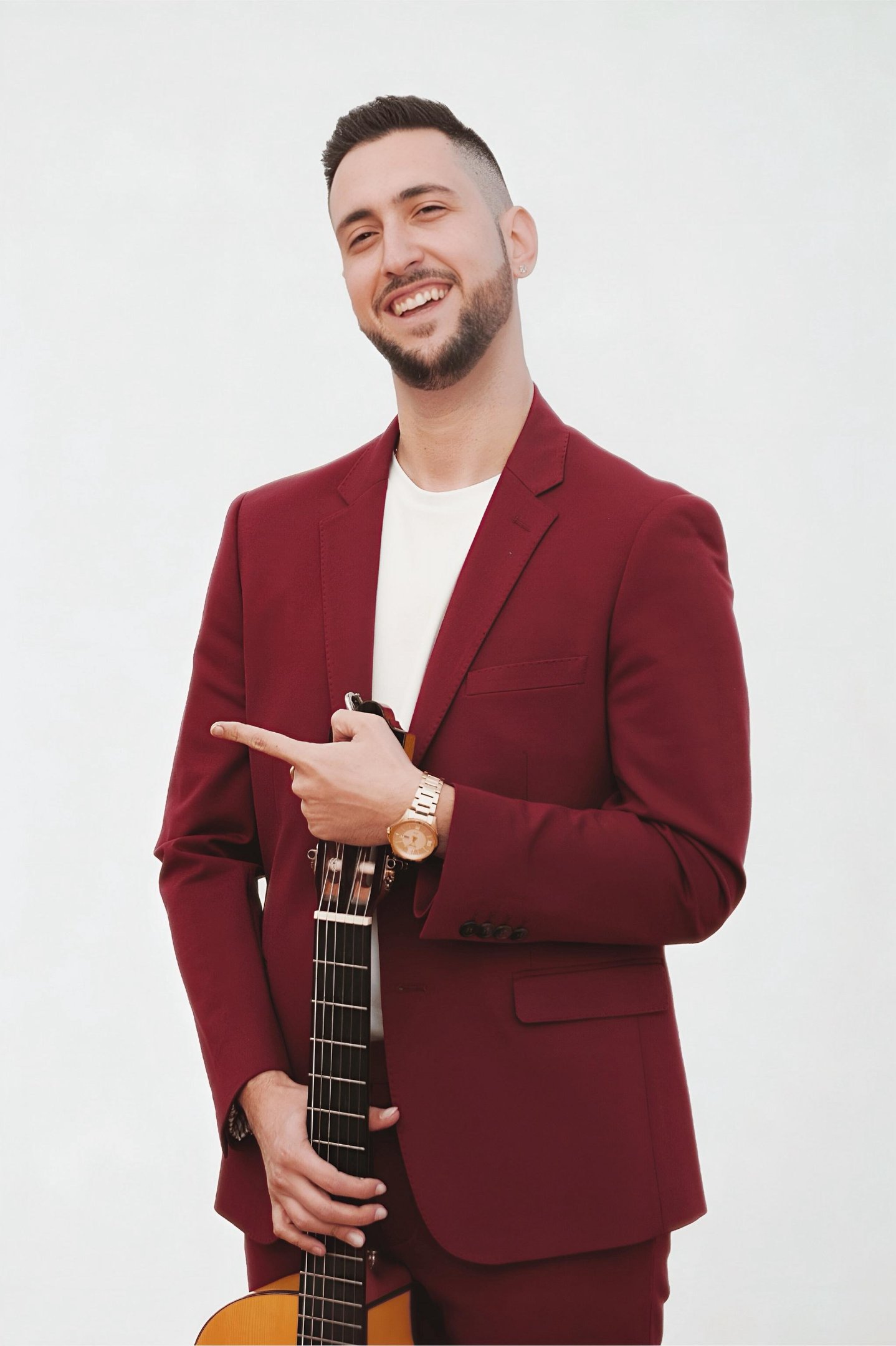 A smiling male musician in a maroon suit holding an acoustic guitar against a white background.