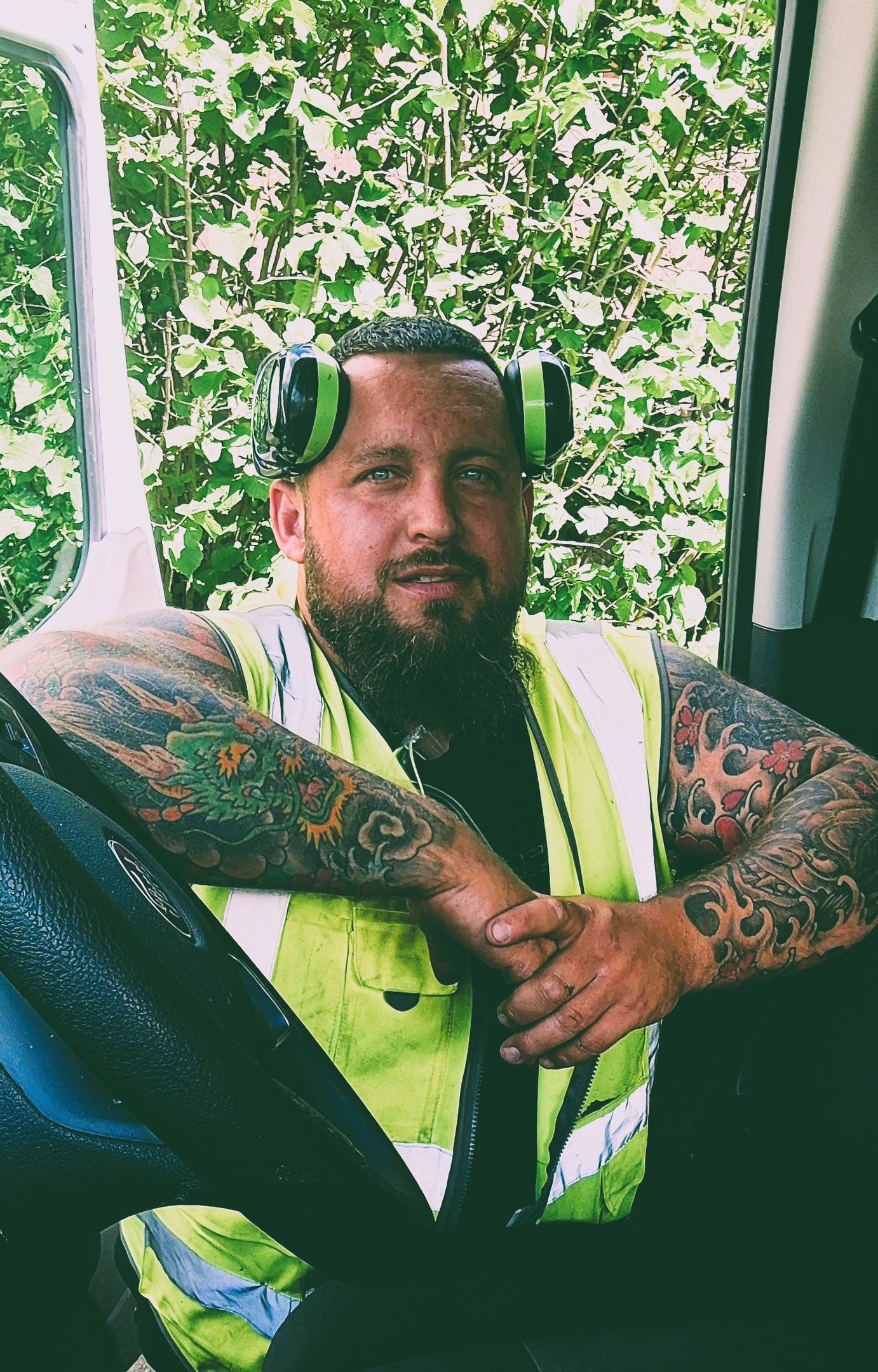 Tattooed construction worker wearing a high-visibility vest and ear protection sitting in a van.