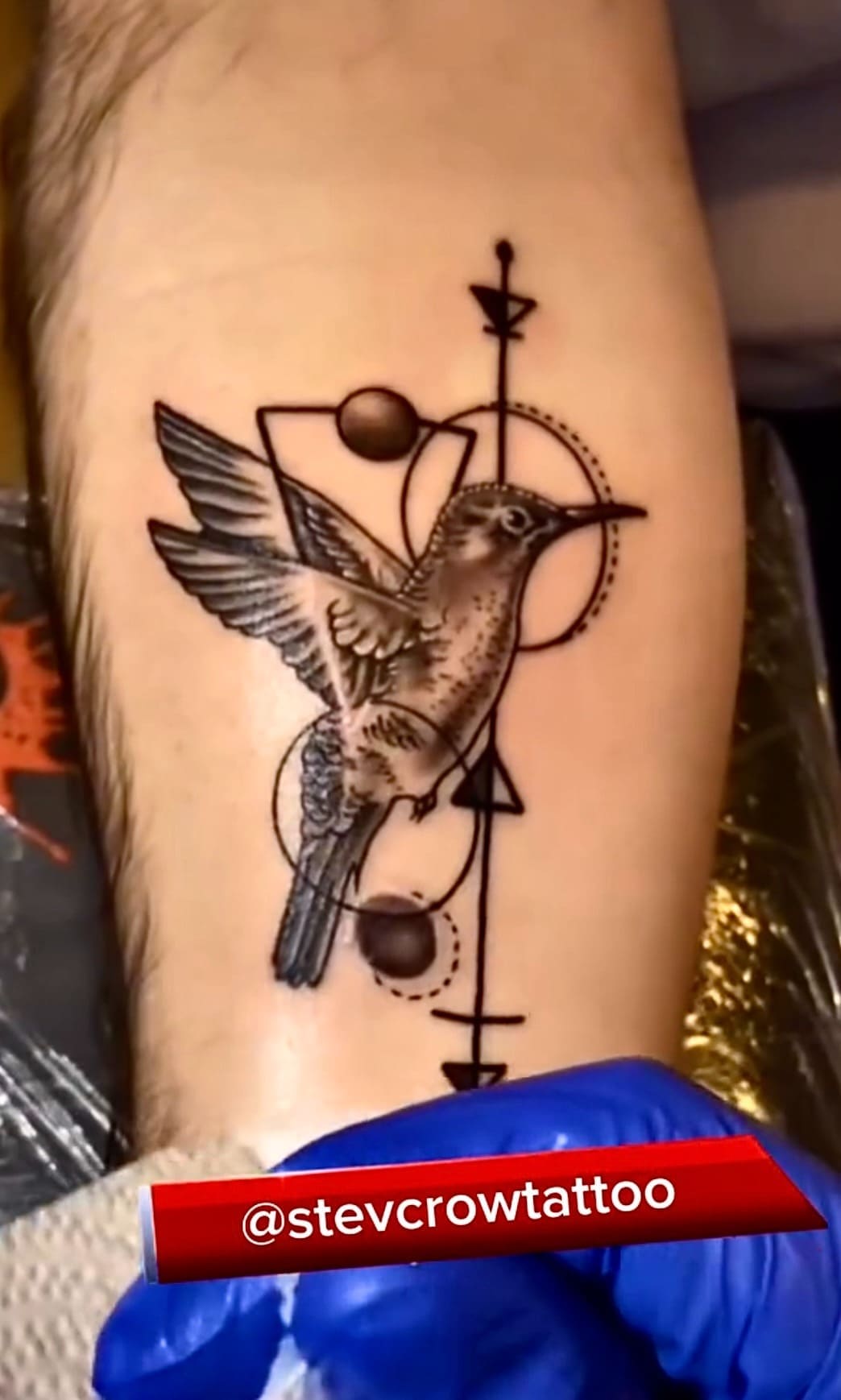 Detailed black and grey hummingbird tattoo with geometric shapes and lines on a forearm.