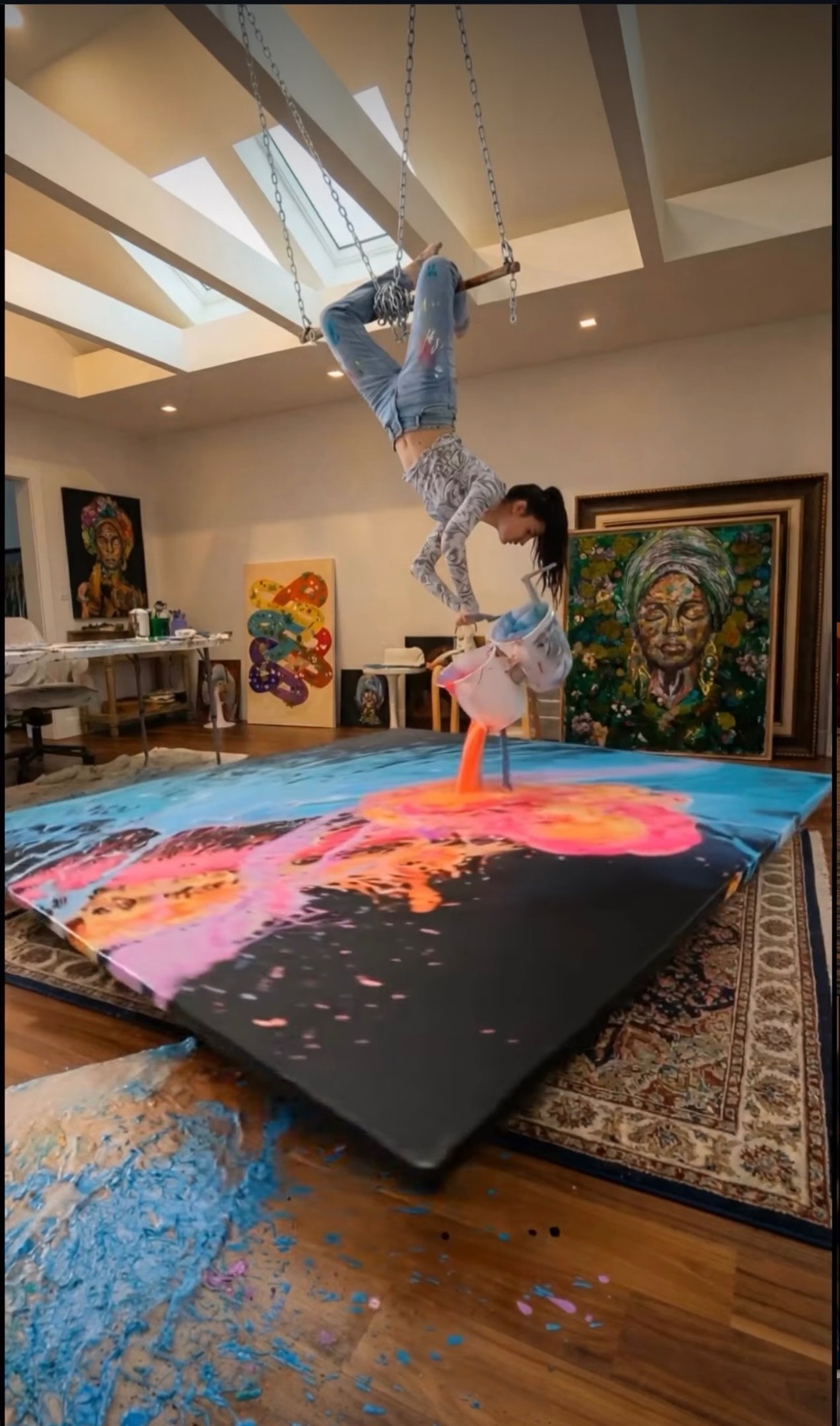 Aerial artist pours vibrant acrylic paint onto a large canvas while hanging upside down in an art studio.