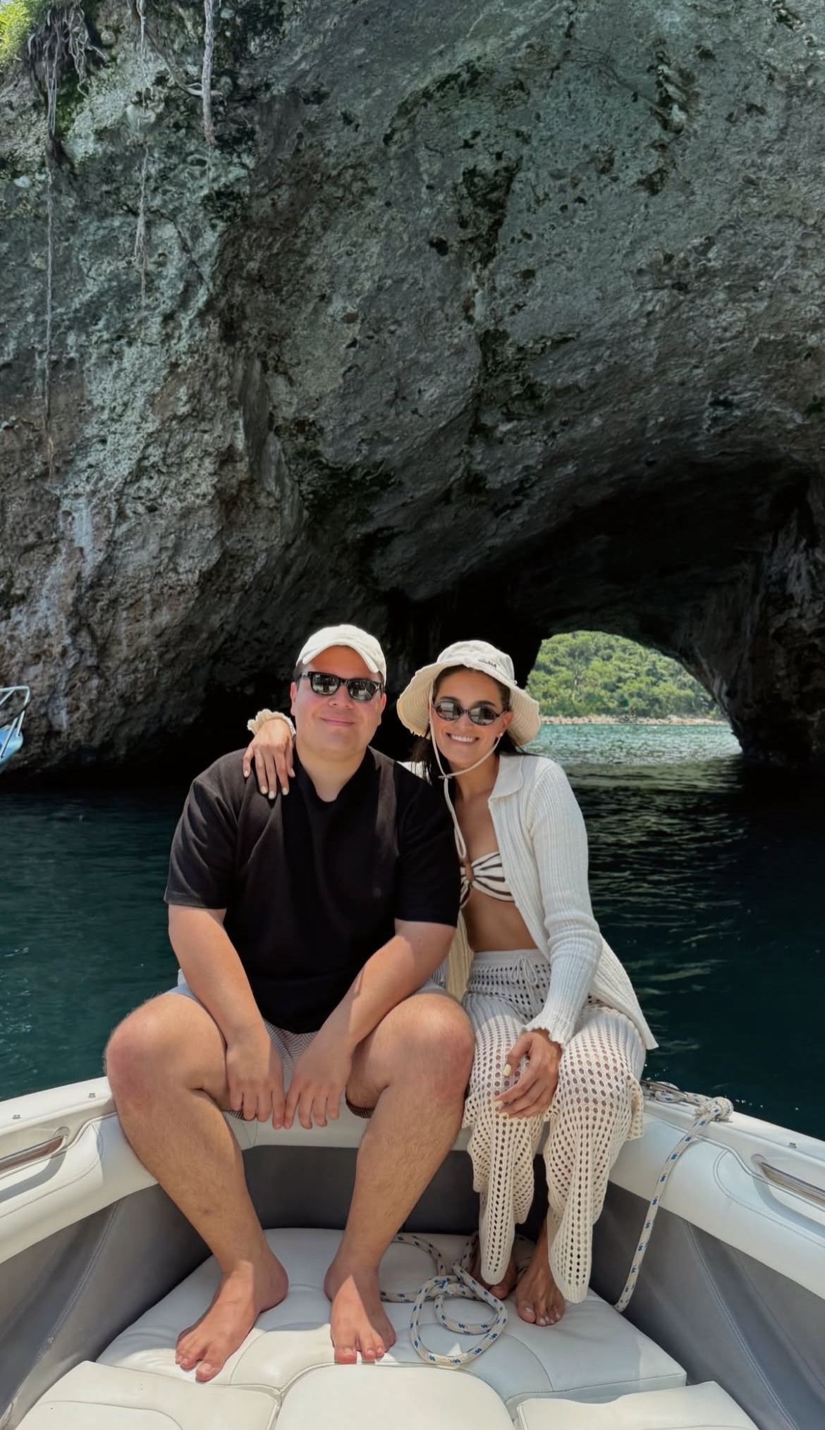 A couple sits on a boat in front of a sea cave archway during a summer vacation boat tour.