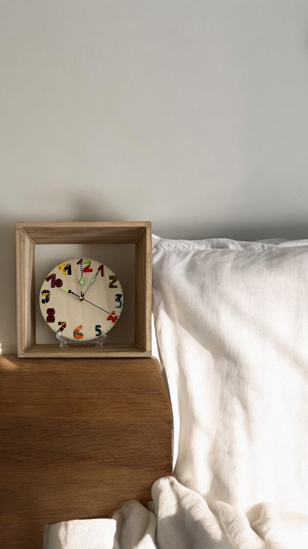 Handmade wooden clock with colorful numbers on a bedside table next to a white pillow.