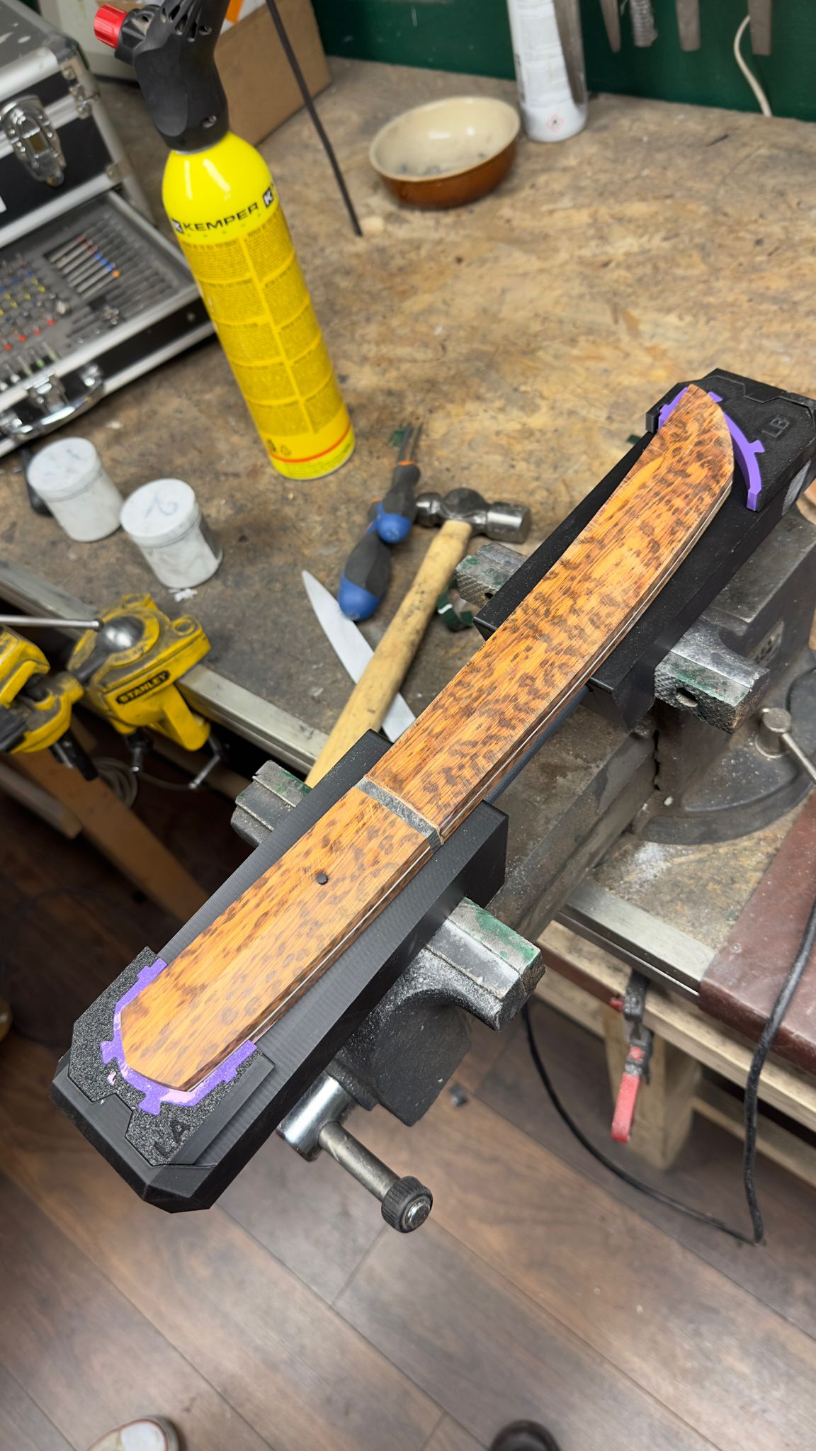Wood grain knife handle clamped in a metal vice on a workshop workbench for crafting.