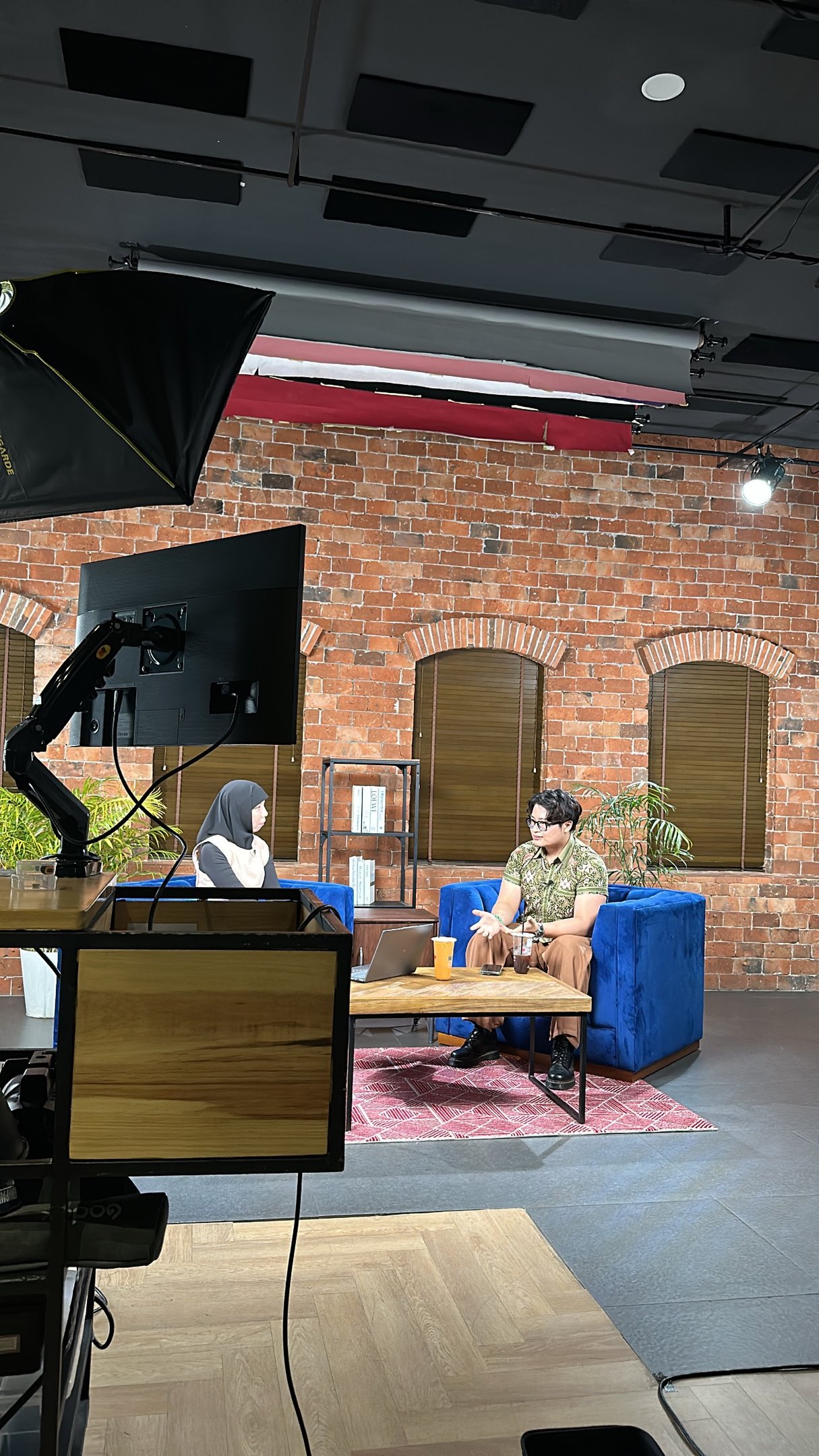Professional podcast studio set with guests on blue velvet sofas against a brick wall background.