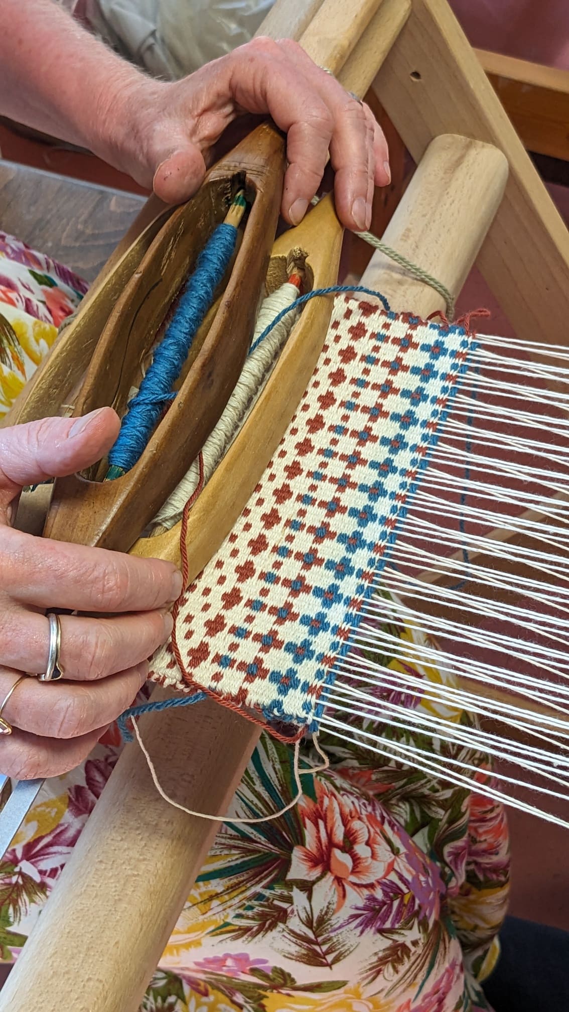 Wooden shuttles and thread spools are used to weave blue, brown, and white crosses on a weaving loom
