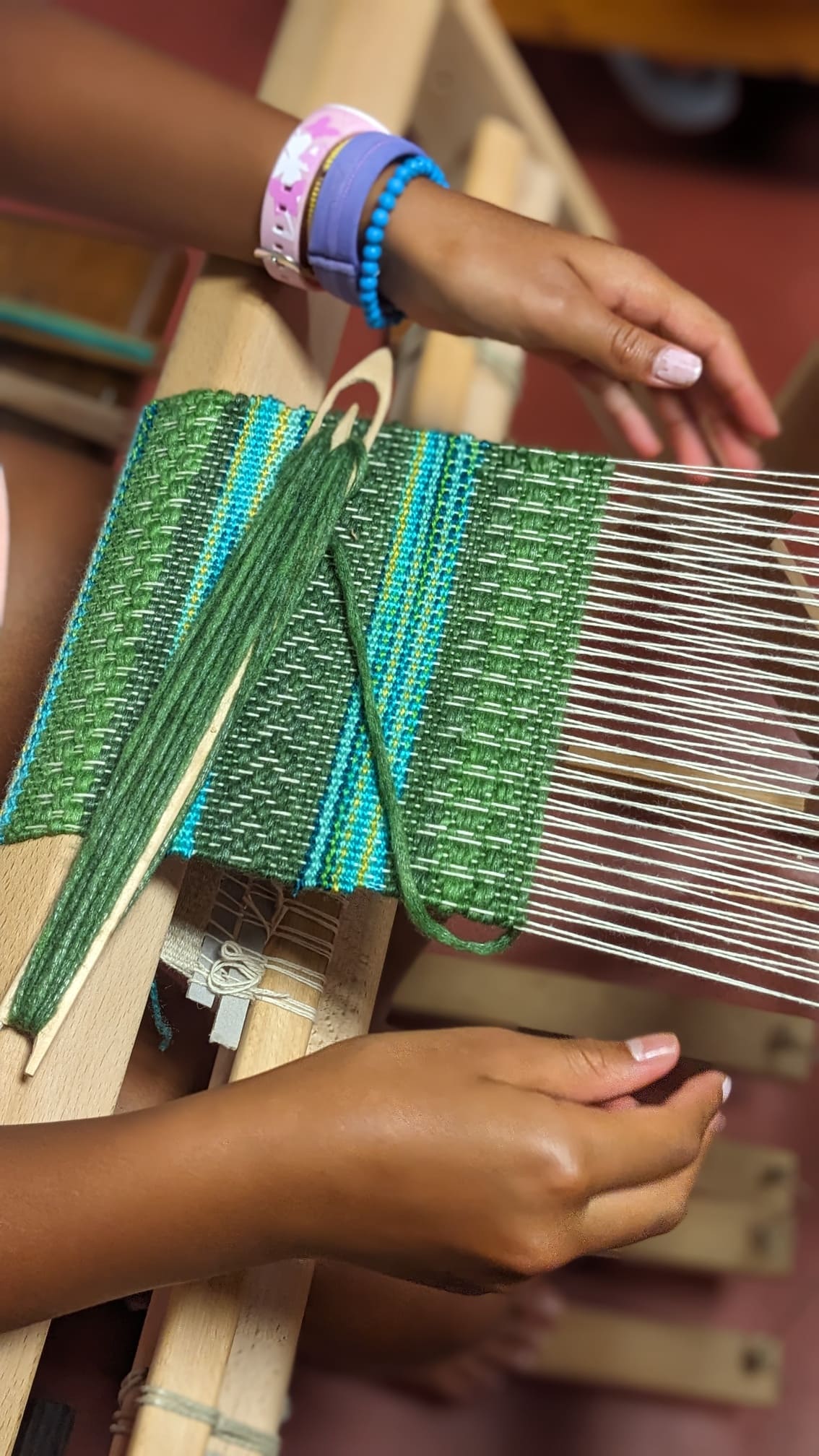 a woman working on a weaving loom with shuttle and green blue threads on a textile in progress