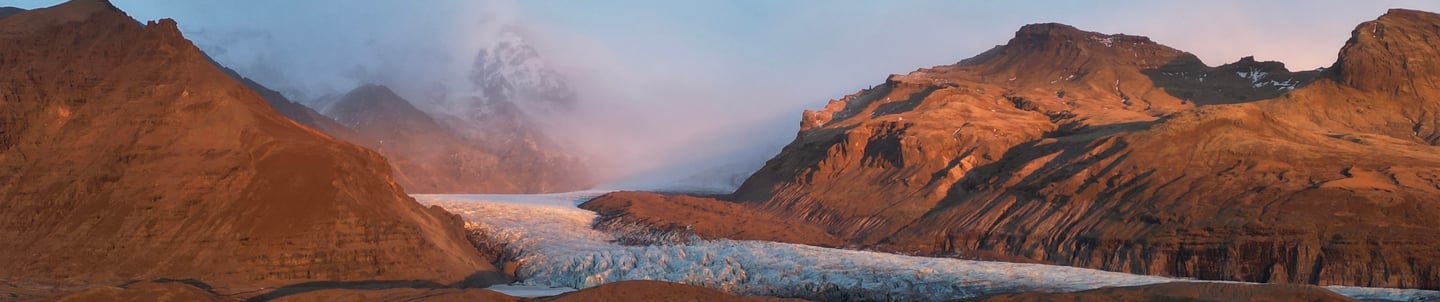Panoramic sunset view of a massive glacier flowing between rugged volcanic mountains in Iceland.