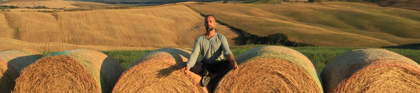 Life coach Abhisek sitting on a hay bale in the sunshine, wuth rolling hills behind him. happiness