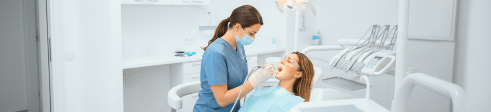 Una dentista con mascarilla y uniforme azul examinando la boca de una paciente 