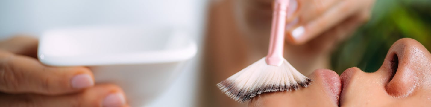 Esthetician applying a professional chemical peel facial treatment to a client using a fan brush.