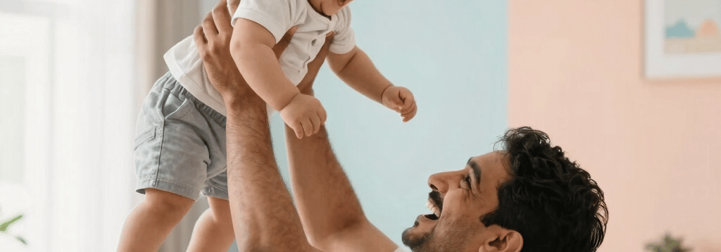 A joyful South Asian father holding his toddler high in the air in a brightly lit modern living room, both laughing. The scene is full of love and trust, soft focus, bright airy pastel colors of peach and light blue in the background.
