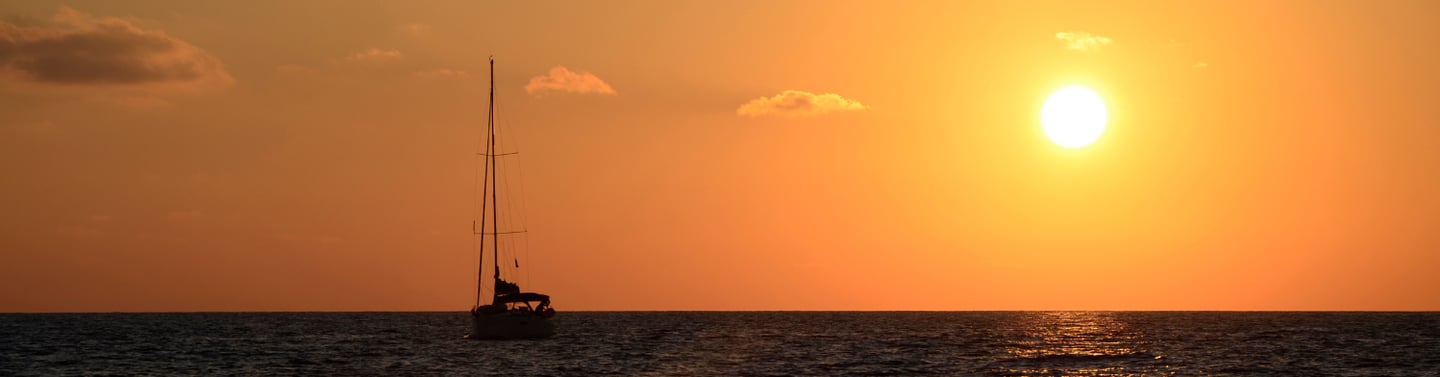 Horizonte durante el atardecer, con el cielo naranja, y el sol y un velero de fondo en el mar