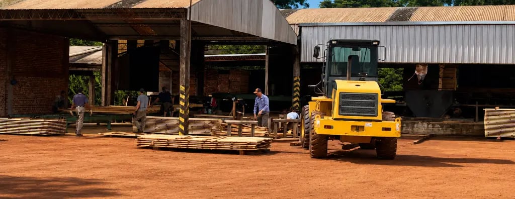 Workers at a lumber mill yard processing wood planks near a yellow front loader tractor.