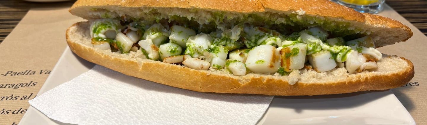 Grilled cuttlefish sandwich with green parsley garlic oil served on a baguette with chips and olives.