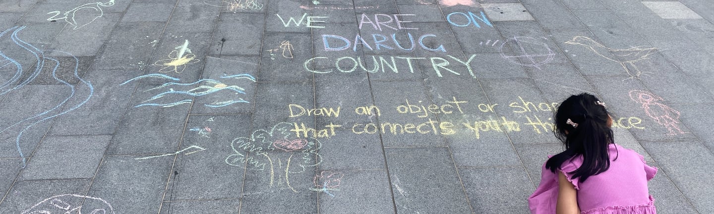 A child with long black hair and a pink dress draws with chalk on pavers. 