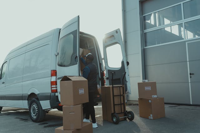 A delivery driver loading cardboard shipping boxes into a white cargo van outside a warehouse.