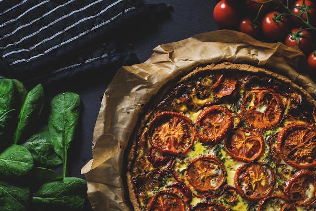 Homemade tomato and spinach quiche on baking paper with fresh ingredients on a dark surface.