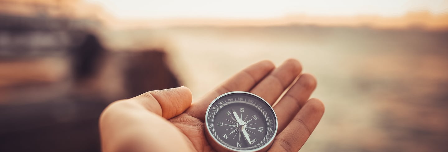 Hand holding a silver pocket compass against a blurred sunset horizon to find his way.