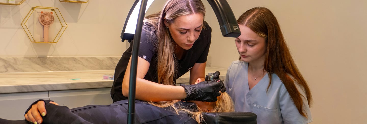 Profesional de microblading enseñando la técnica a alumna durante práctica en modelo con aro de luz en cabina blanca y beige.