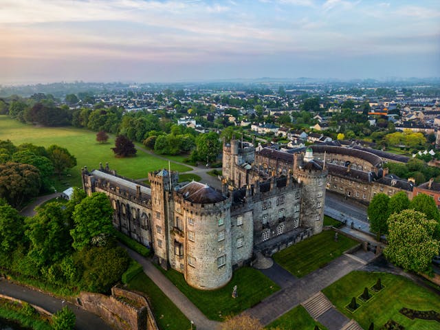 Aerial view of Kilkenny Castle in Ireland surrounded by lush green gardens and a historic city skyline.