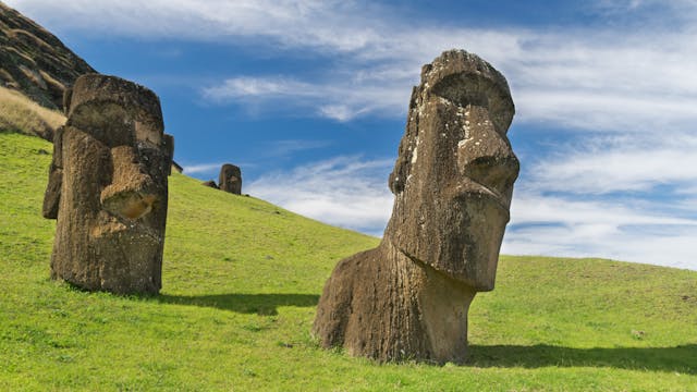 Ancient Moai stone statues on a grassy hill at Easter Island under a blue sky.