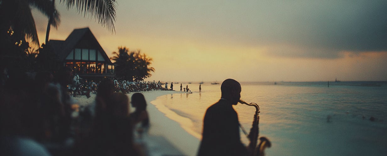 A silhouetted musician plays the saxophone on a tropical beach at sunset during a seaside party.