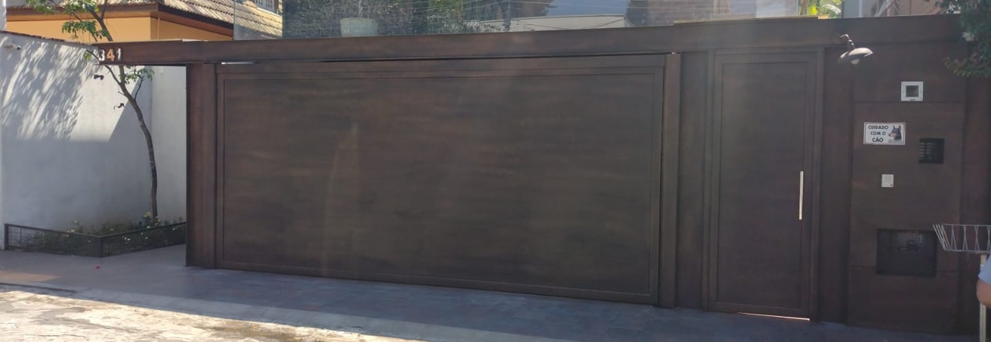 Vista aproximada do portão de garagem basculante e do portão social com acabamento em pintura que imita o aço corten, em fren