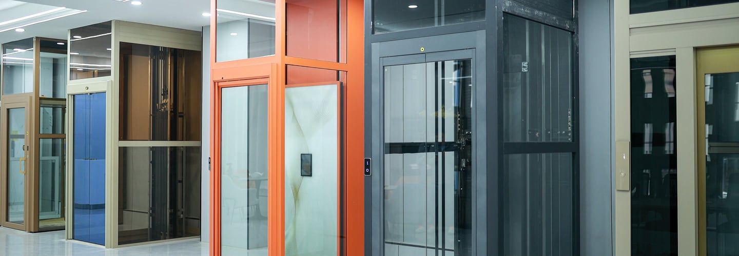 Modern glass home elevator models in a showroom featuring orange and black frames for residential accessibility.