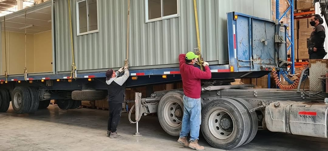 Logistics workers securing a modular portable shipping container office onto a semi-truck trailer for transport.