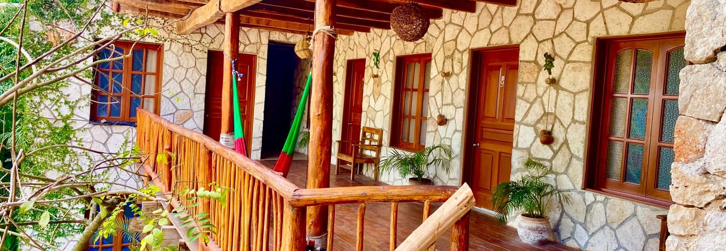 Rustic stone villa balcony with wooden beams, hanging hammocks, and tropical plants overlooking a pool.