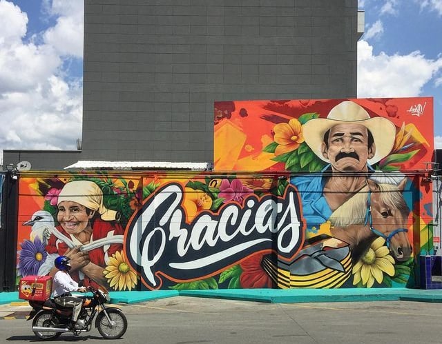 Vibrant outdoor street art mural featuring Latin American figures, flowers, and the word Gracias.