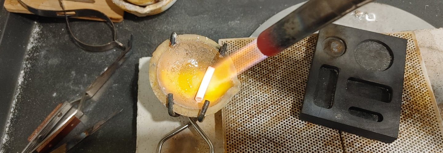 A silver jewelry-making scene showing a crucible held by tongs, heated by a torch.  