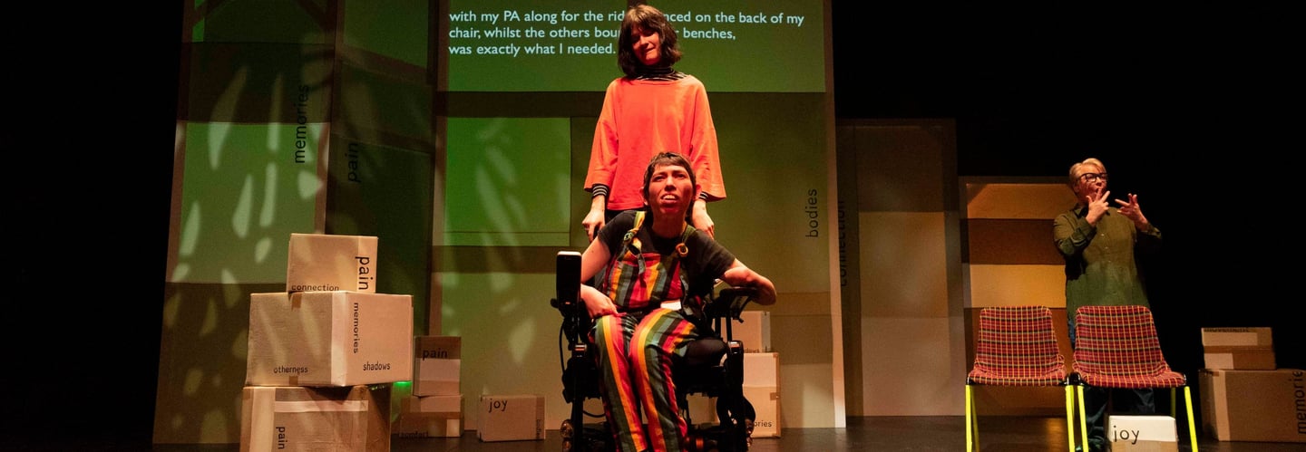 A white electric wheelchair user in front of a white person in orange. A BSL interpreter to the side