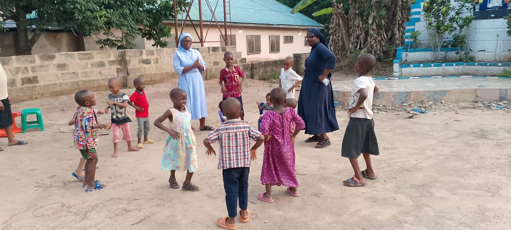 Two Catholic nuns playing outdoors with a group of young African children in a village courtyard.
