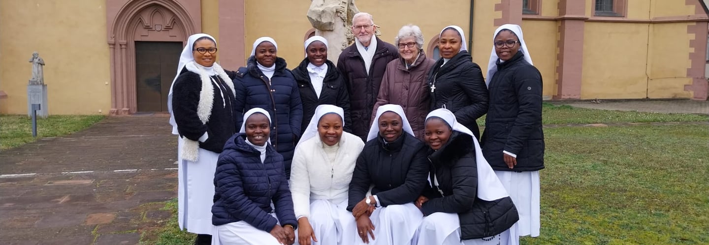 A group of Catholic nuns in white habits and winter coats posing with two seniors outside a church.