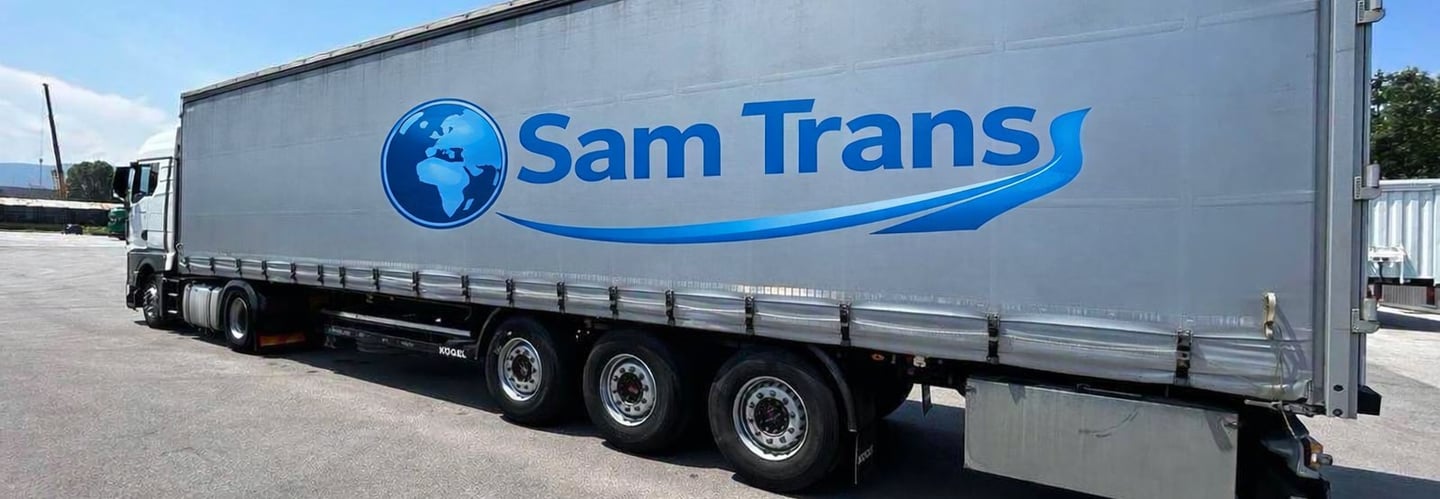 A silver Sam Trans logistics semi-truck trailer parked on asphalt under a clear blue sky.