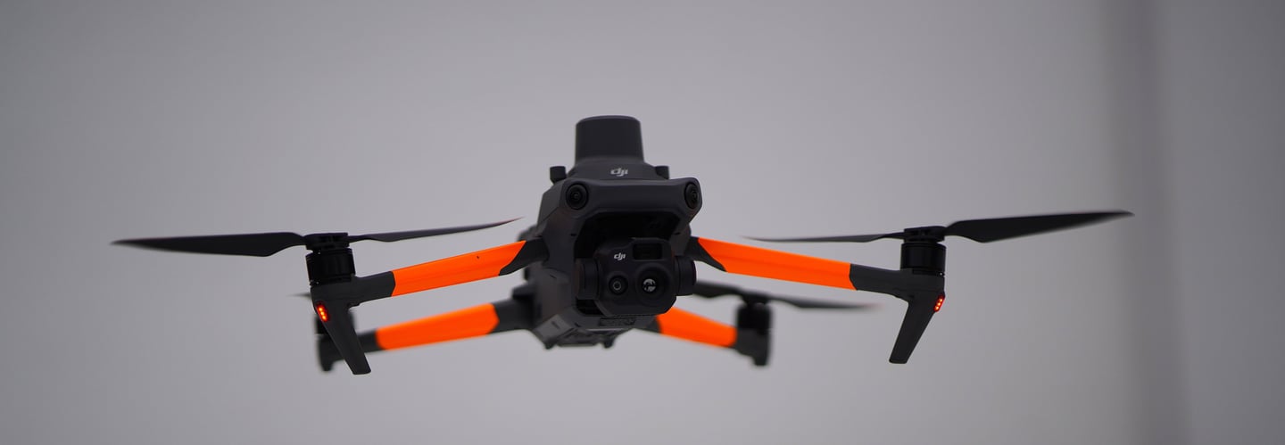 A DJI Mavic 3 Enterprise thermal drone with orange arms hovering in flight against a grey background.